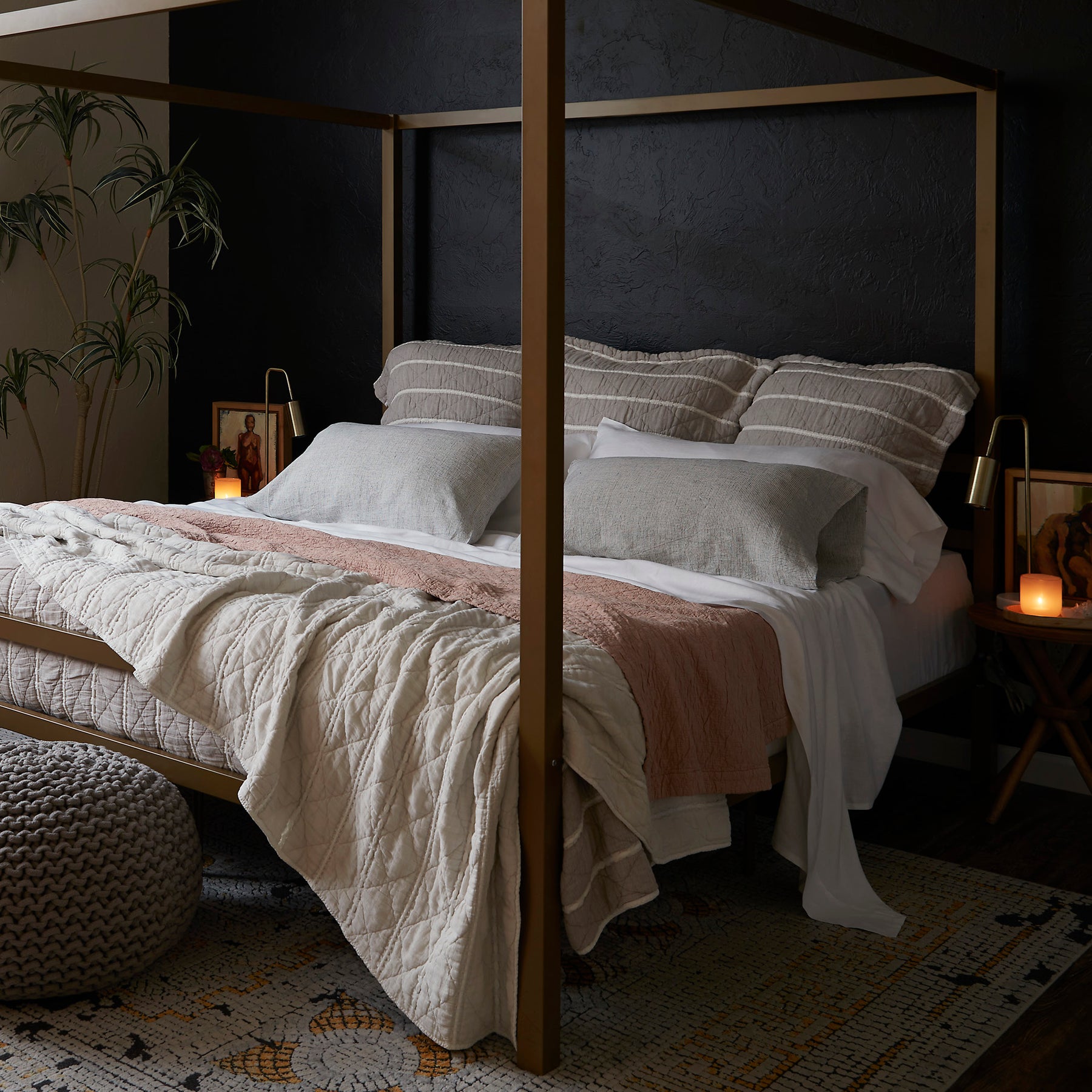 Image of a made bed with a dark background. The bed showcases pillows of various sizes and layered bedding. Items on the bed include: Kapok Pillows, White Blended Linen Sheets, Pinstripe Relaxed Hemp Pillowcases, a Pink Sandstone Wave Coverlet, and a reversible gray and white striped Heritage Quilt.