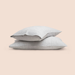 Image of two pillows with Pinstripe Relaxed Hemp pillowcases stacked on top of each other on a light pink background. The top pillow is showcasing an enveloping feature. 