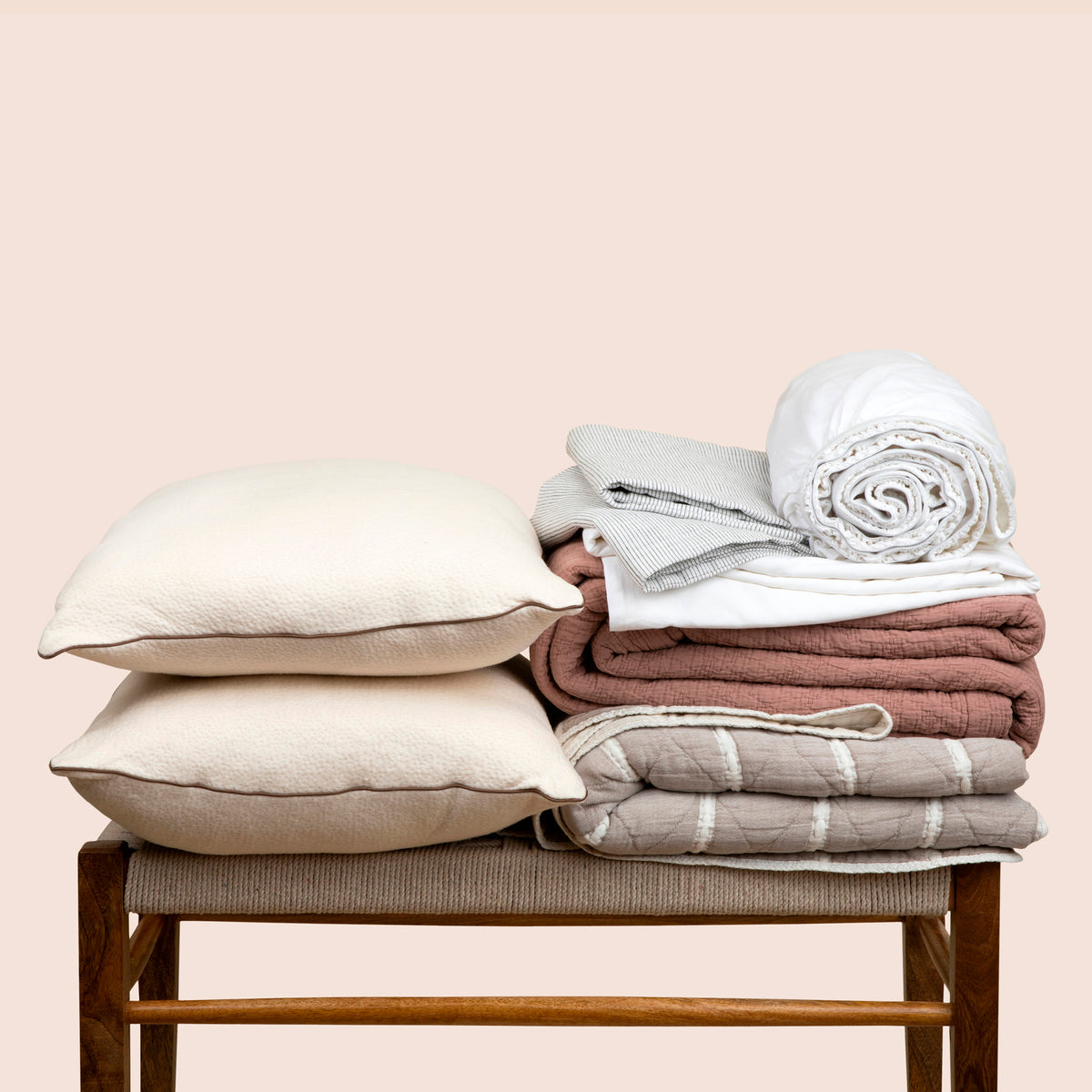 Image showcasing the complete Heritage Bundle neatly folded on a bench. The set showcases: two Kapok pillows stacked on each other on the left side of the bench and a stack including: White Blended Linen Sheets, Pinstripe Relaxed Hemp Pillowcases, a Pink Sandstone Wave Coverlet, and a reversible gray and white striped Heritage Quilt on the right side. 