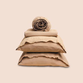 Image showcasing entire Ochre Garment Washed Percale Sheet Set on a light pink background. Set features a rolled-up fitted sheet on top, a neatly folded flat sheet, and two stacked pillowcases. 