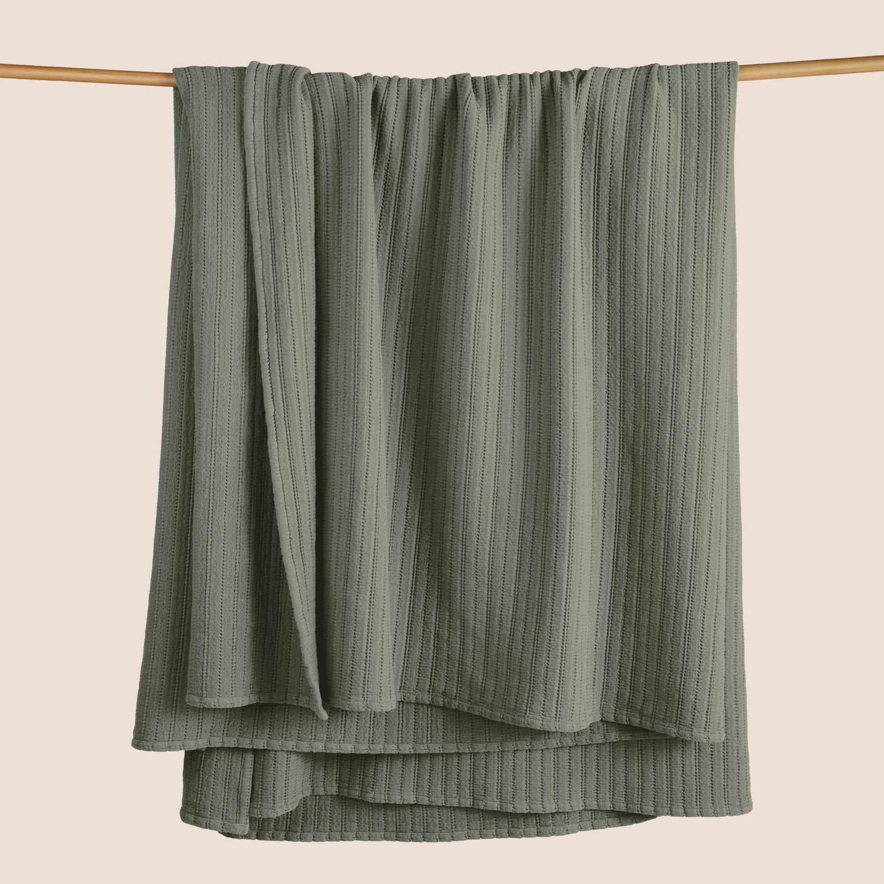 Image of an Agave Ridgeback Coverlet hanging and draped over a wooden rod on a light pink background