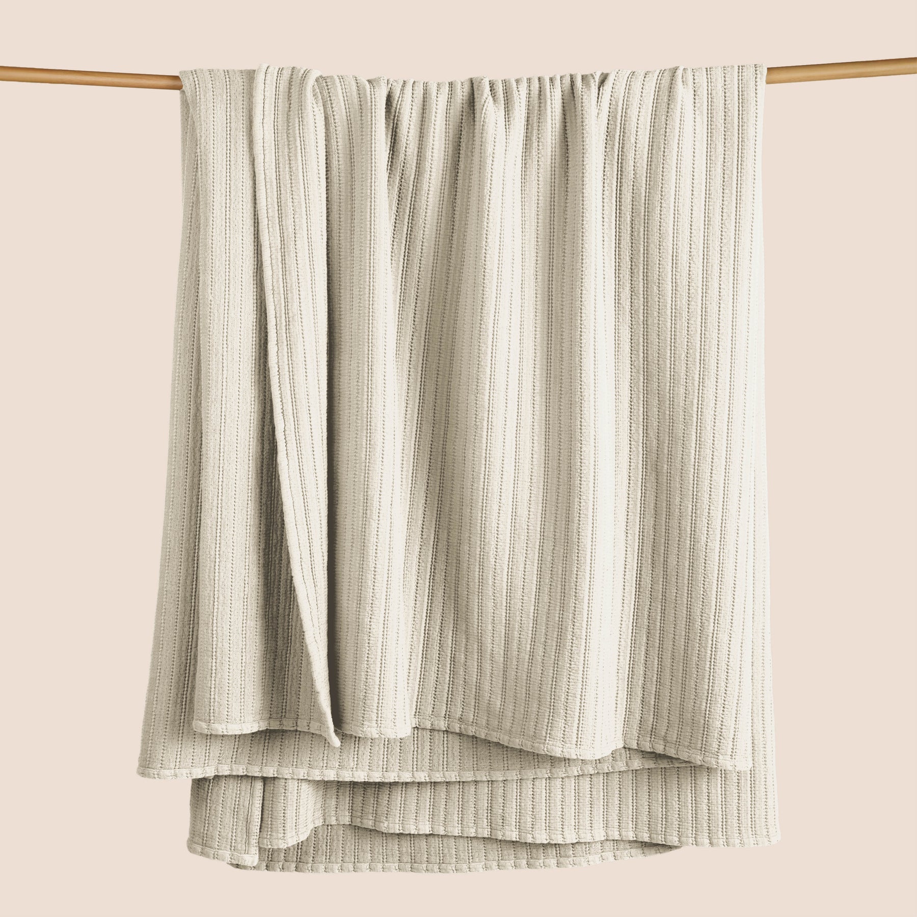 Image of an Ecru Ridgeback Coverlet hanging and draped over a wooden rod on a light pink background