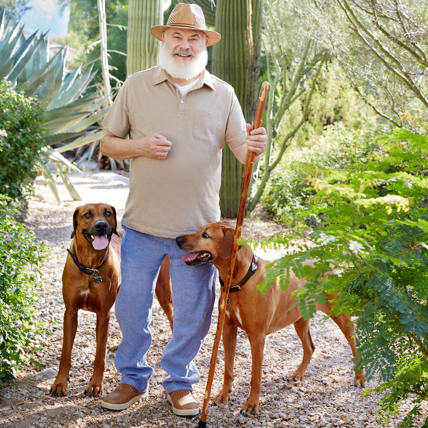 Image of Dr. Andrew Weil standing outside on a gravel path between various plants. There are two Rhodesian Ridgeback dogs standing on either side of him.
