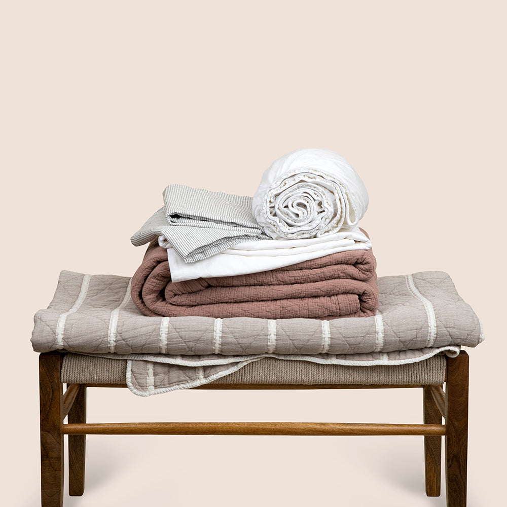 Image showcasing the Heritage Bundle, featuring the a brown bench with folded bedding on top. From bottom to top: gray and white Heritage Quilt, Pink Sandstone Wave Coverlet, White Blended Linen Sheets, Pinstripe Relaxed Hemp Pillowcases.