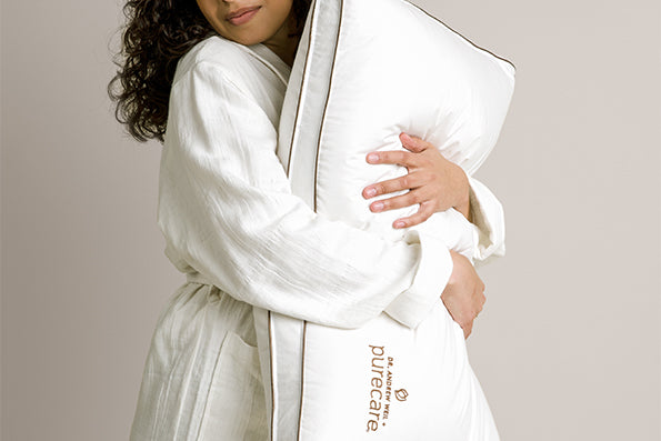 Image of woman in a white robe, softly smiling and hugging a Chambered Down Pillow close to her 