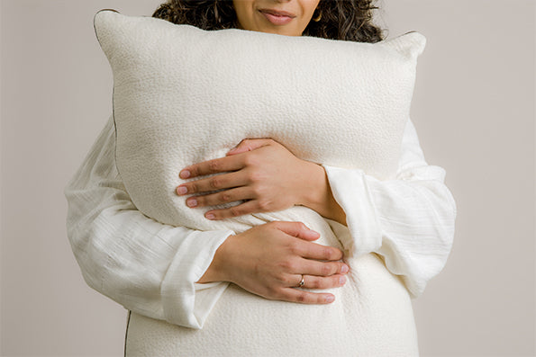 Image of a woman softly smiling in a white robe, holding and hugging the Perfect Kapok Pillow