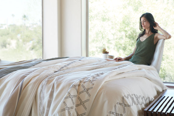 Image of a woman in green pajamas sitting in a chair by her bed in a naturally-lit room. The bed features a Sonoran Duvet Cover on it.