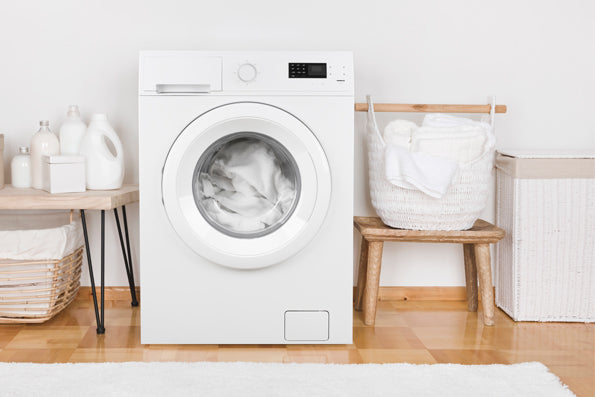 Image of a white washing machine with white sheets inside. The room is filled with various white and light wood decor and furniture 