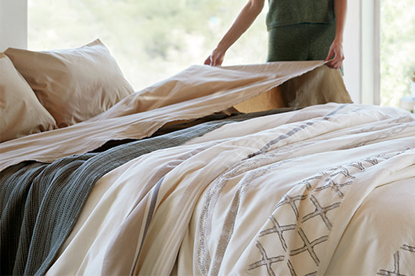 Image of a woman in a green shirt making her bed in a naturally-lit room. The bed includes Ochre Garment Washed Percale Sheets, an Agave Ridgeback Coverlet, and a Sonoran Duvet Cover.