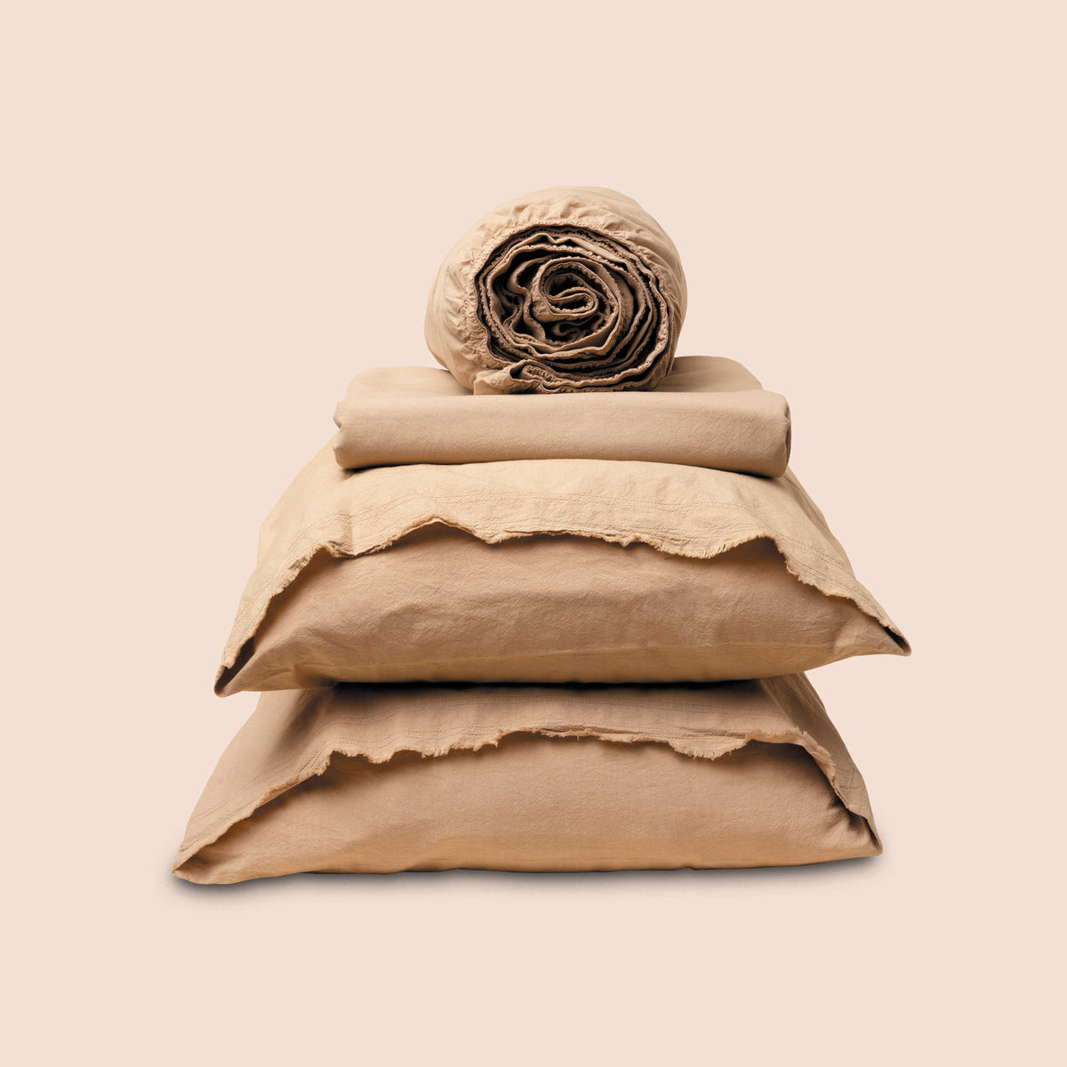 Image showcasing entire Ochre Garment Washed Percale Sheet Set on a light pink background. Set features a rolled-up fitted sheet on top, a neatly folded flat sheet, and two stacked pillowcases with raw-edged detailing facing front. 
