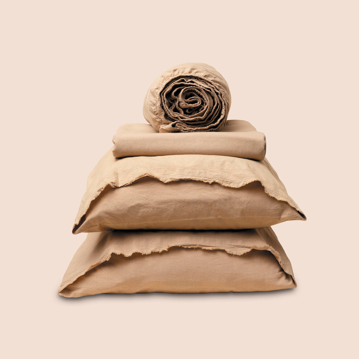 Image showcasing entire Ochre Garment Washed Percale Sheet Set on a light pink background. Set features a rolled-up fitted sheet on top, a neatly folded flat sheet, and two stacked pillowcases with raw-edged detailing facing front.