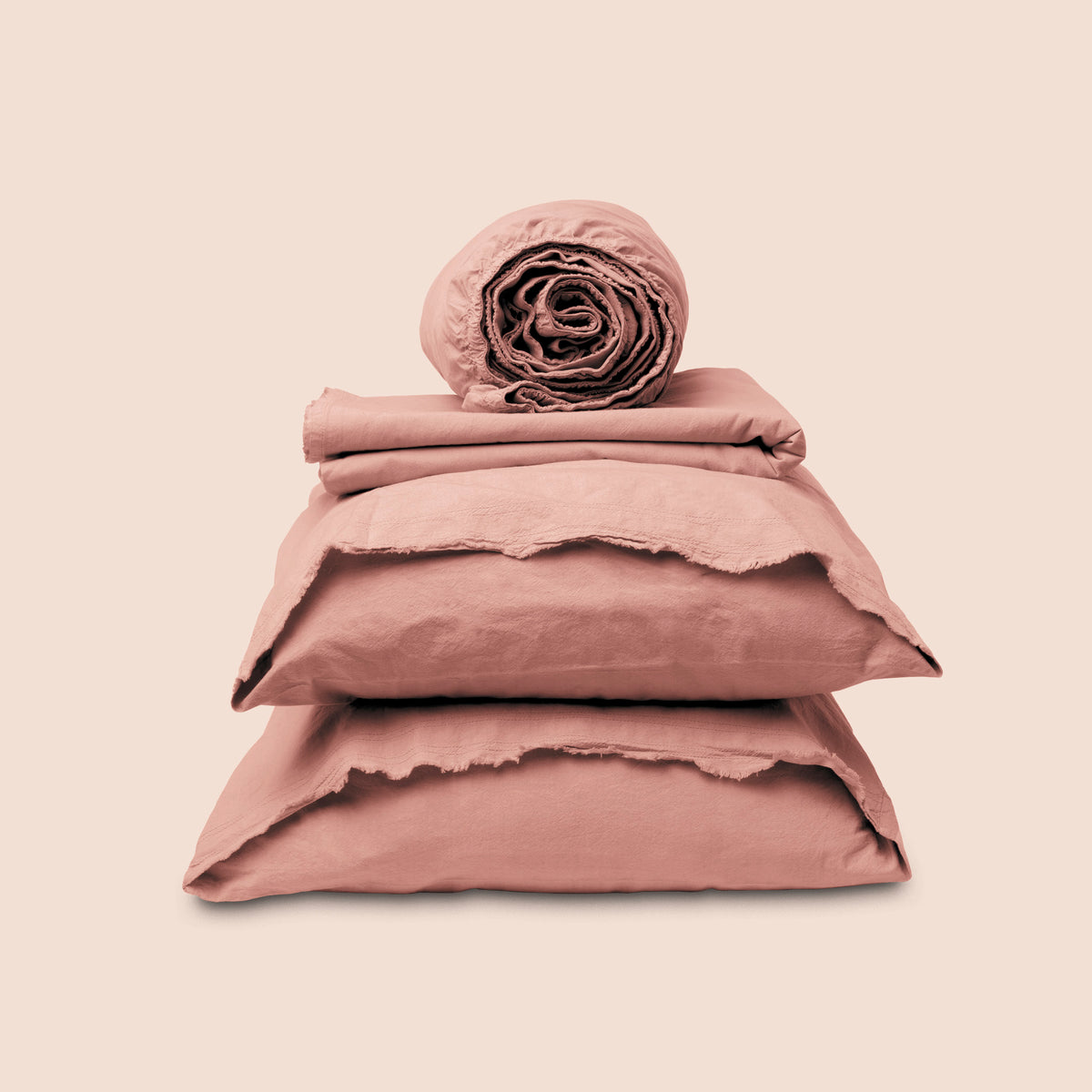 Image showcasing entire Pink Sandstone Garment Washed Percale Sheet Set on a light pink background. Set features a rolled-up fitted sheet on top, a neatly folded flat sheet, and two stacked pillowcases with raw-edged detailing facing front. 