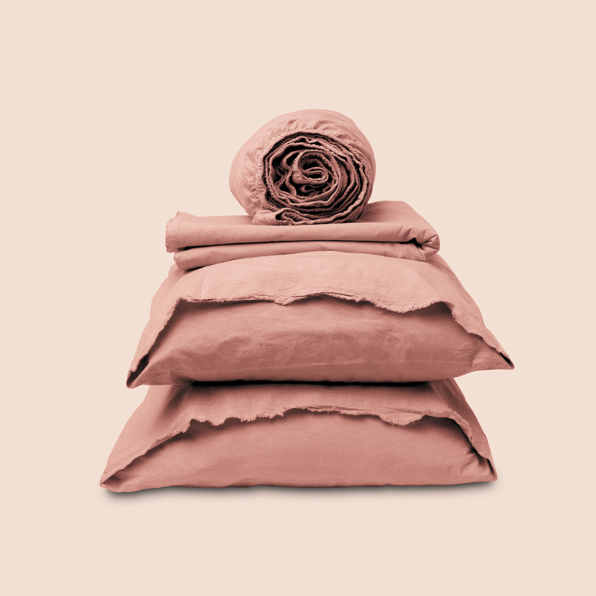 Image showcasing entire Pink Sandstone Garment Washed Percale Sheet Set on a light pink background. Set features a rolled-up fitted sheet on top, a neatly folded flat sheet, and two stacked pillowcases with raw-edged detailing facing front.