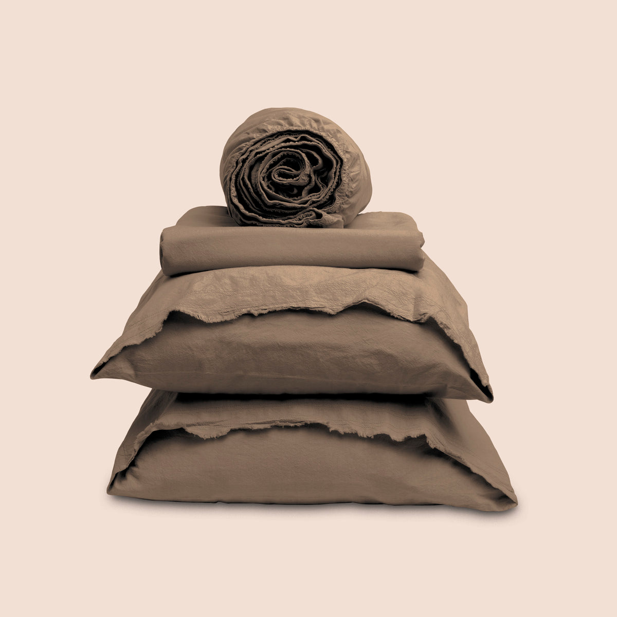 Image showcasing entire Desert Sand Garment Washed Percale Sheet Set on a light pink background. Set features a rolled-up fitted sheet on top, a neatly folded flat sheet, and two stacked pillowcases with raw-edged detailing facing front. 