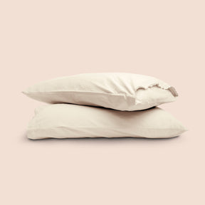 Image of two pillows with Ecru Garment Washed Percale pillowcases stacked on top of each other on a light pink background. The top pillow is showcasing an enveloping feature and raw edge design. 