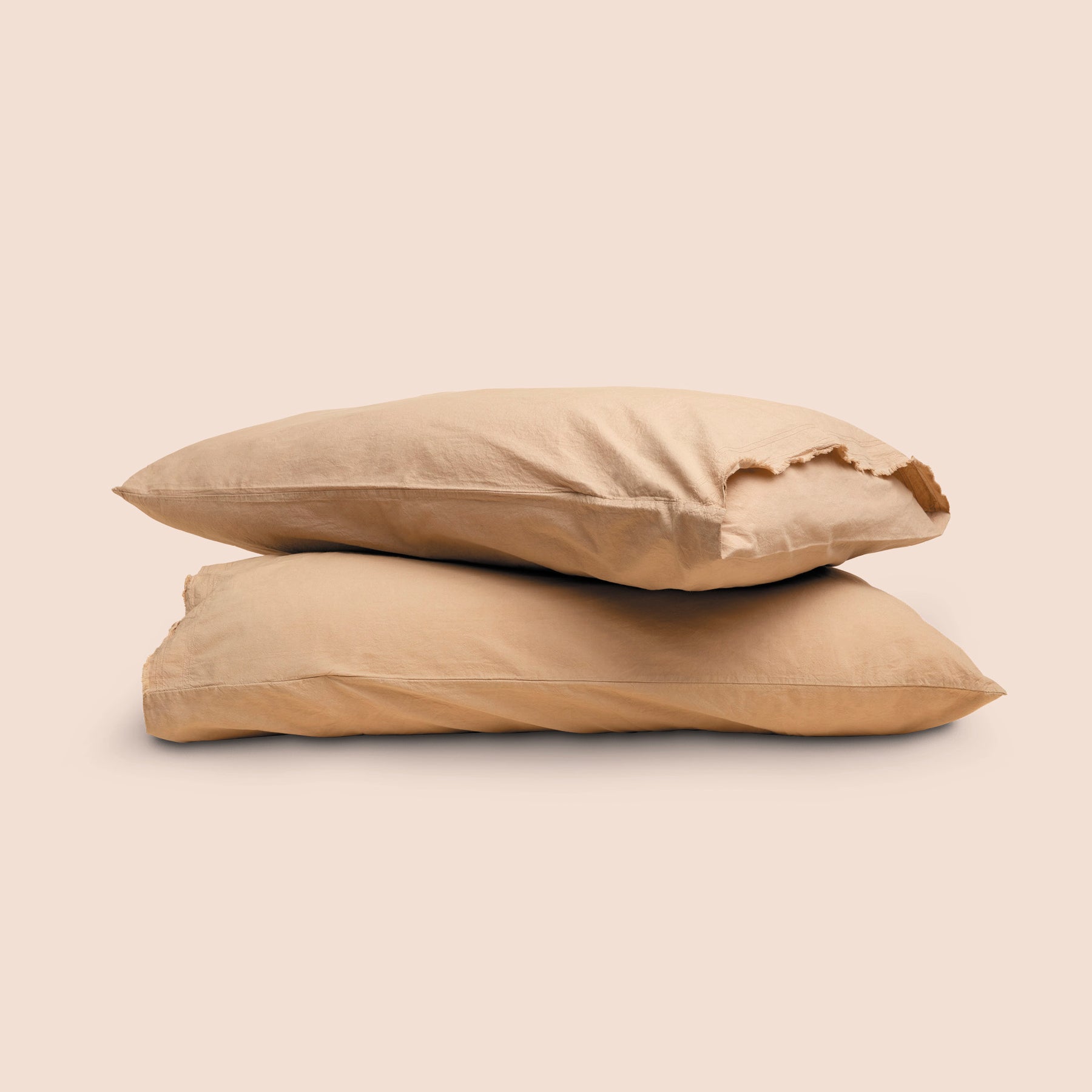 Image of two pillows with Ochre Garment Washed Percale pillowcases stacked on top of each other on a light pink background. The top pillow is showcasing an enveloping feature and raw edge design. 