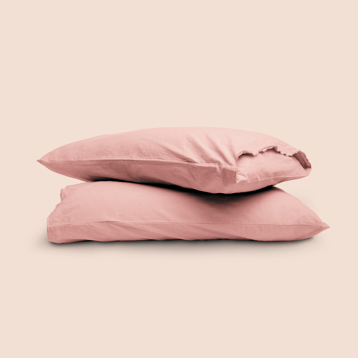 Image of two pillows with Pink Sandstone Garment Washed Percale pillowcases stacked on top of each other on a light pink background. The top pillow is showcasing an enveloping feature. 