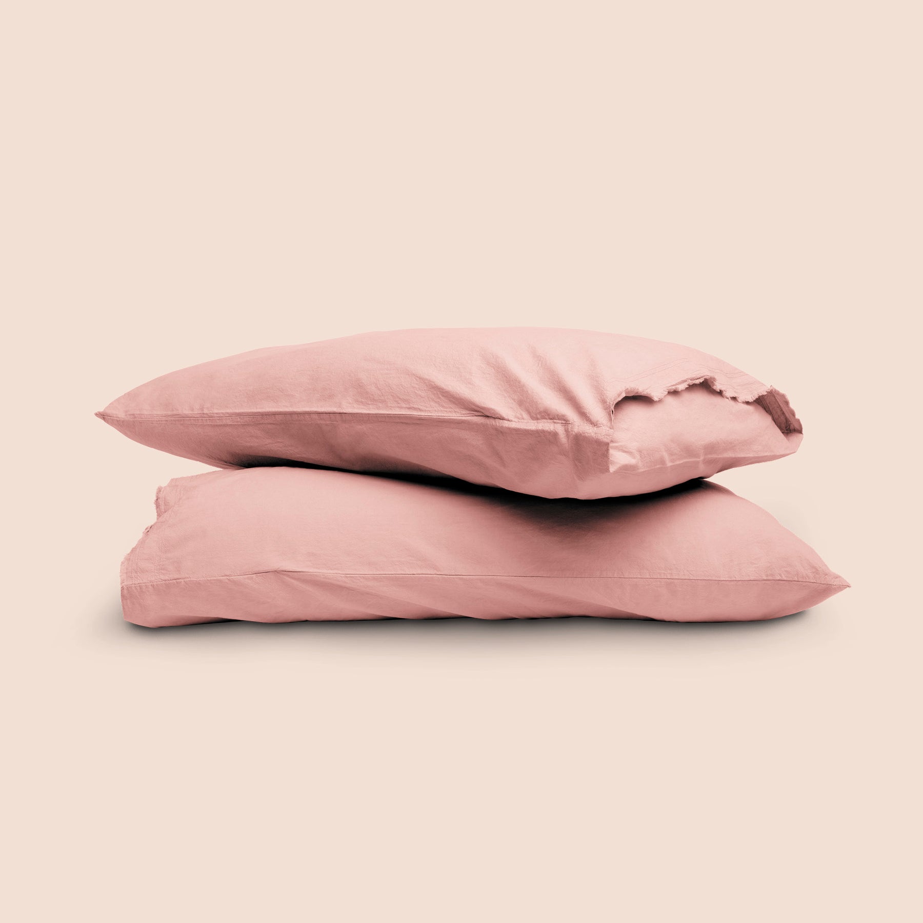 Image of two pillows with Pink Sandstone Garment Washed Percale pillowcases stacked on top of each other on a light pink background. The top pillow is showcasing an enveloping feature and raw edge design. 