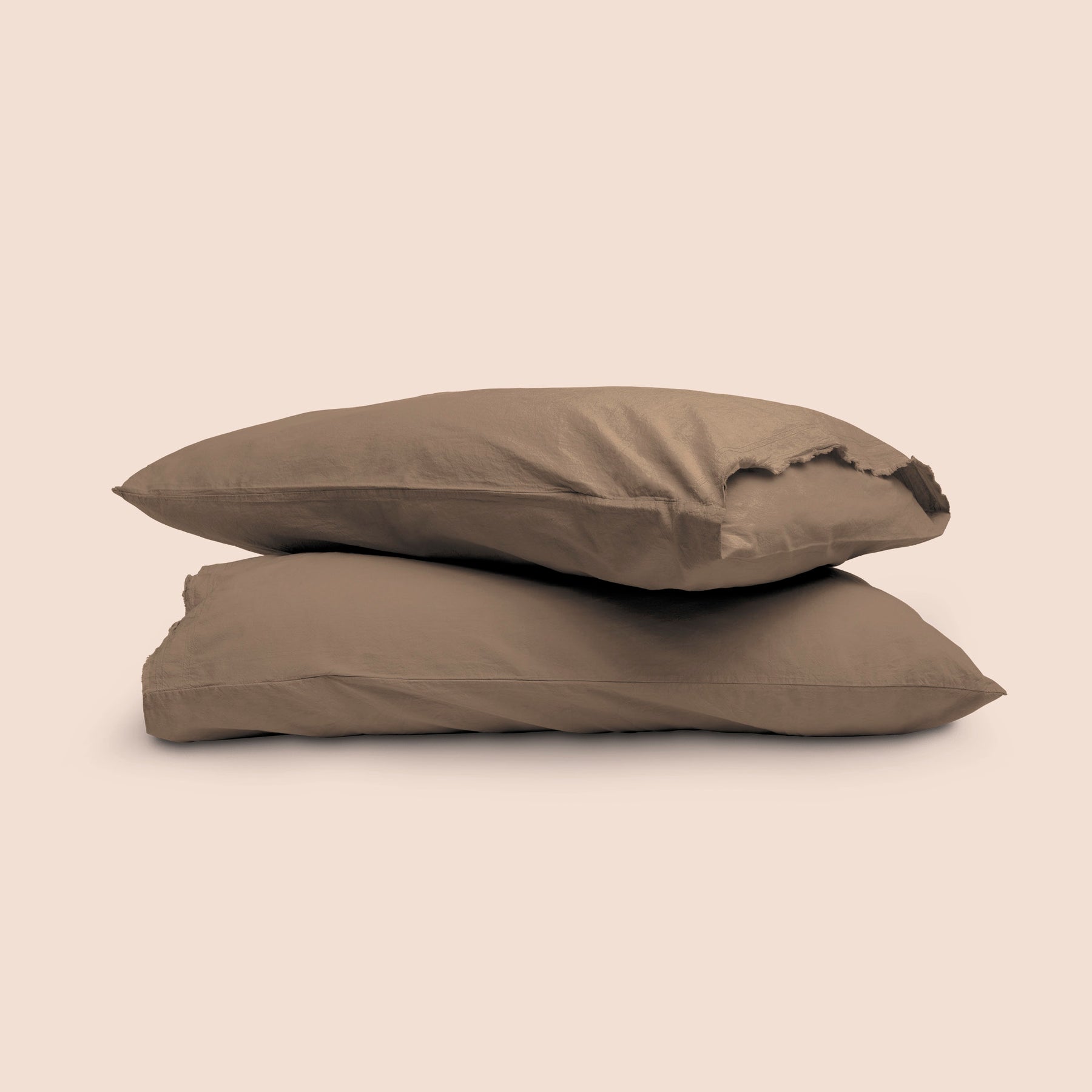 Image of two pillows with Desert Sand Garment Washed Percale pillowcases stacked on top of each other on a light pink background. The top pillow is showcasing an enveloping feature and raw edge design. 