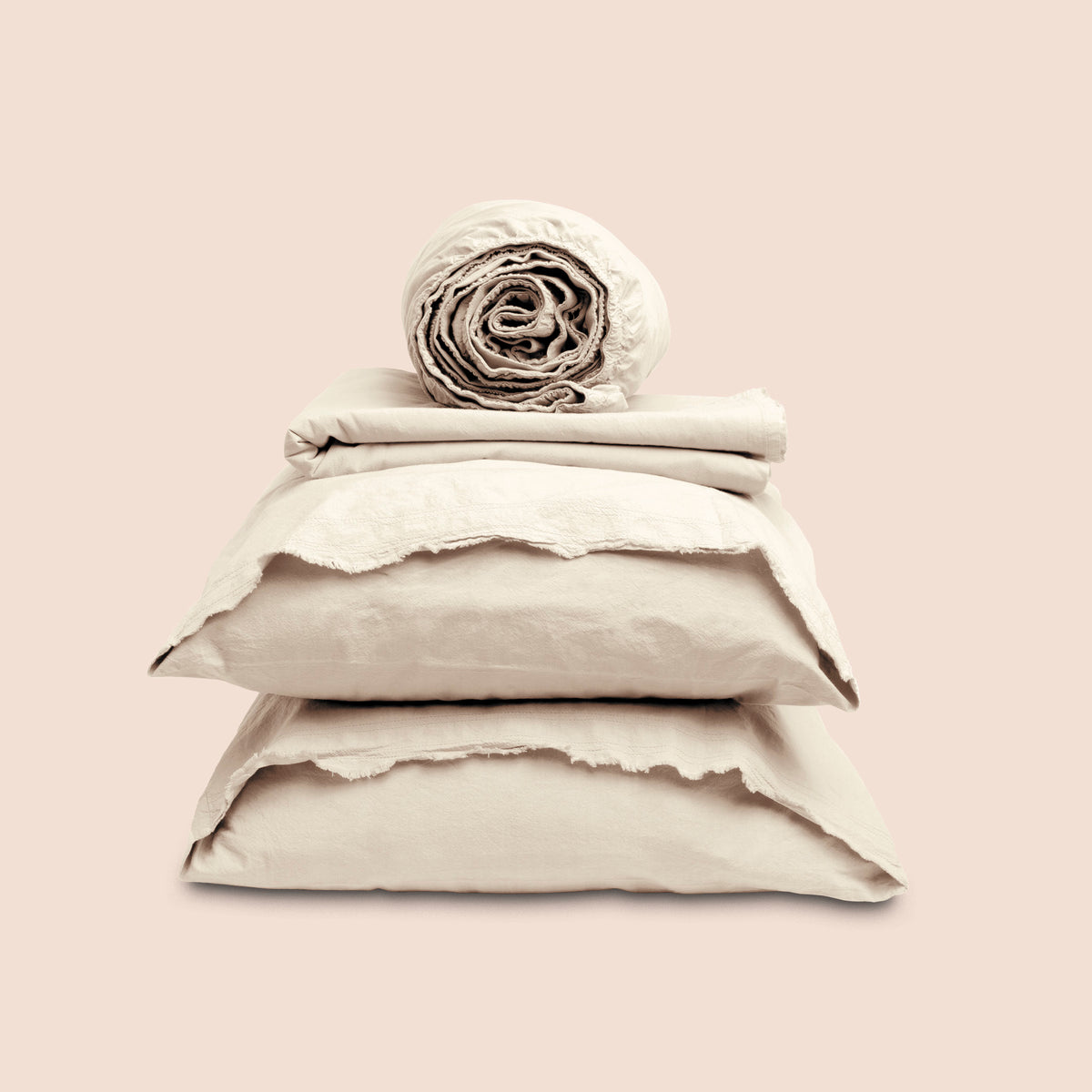 Image showcasing entire Ecru Garment Washed Percale Sheet Set on a light pink background. Set features a rolled-up fitted sheet on top, a neatly folded flat sheet, and two stacked pillowcases with raw-edged detailing facing front. 