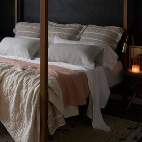 Image of a made bed with a dark background. The bed showcases pillows of various sizes and layered bedding. Items on the bed include: White Blended Linen Sheets, Pinstripe Relaxed Hemp Pillowcases, a Pink Sandstone Wave Coverlet, and a reversible gray and white striped Heritage Quilt.