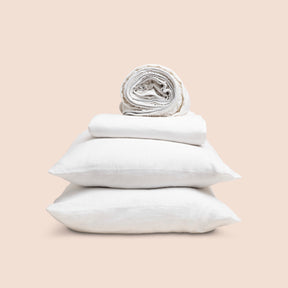 Image showcasing entire White Relaxed Hemp Sheet Set on a light pink background. Set features a rolled-up fitted sheet on top, a neatly folded flat sheet, and two stacked pillowcases. 