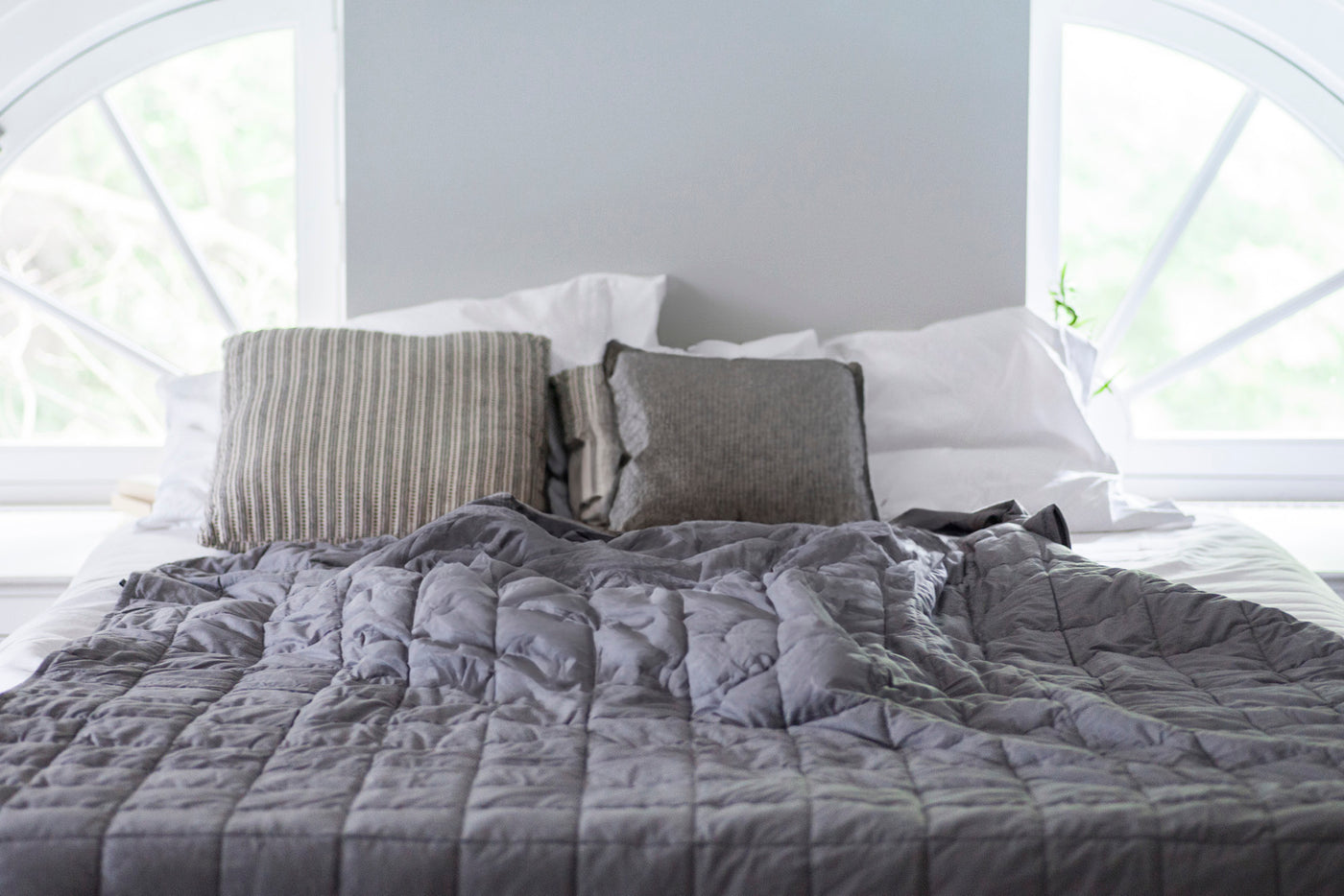 Image of a bed in a bright white room with white sheets and a gray Weighted Blanket on top