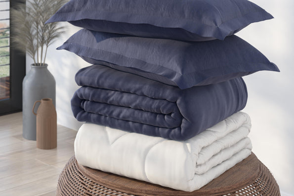 Image of a pile of bedding stacked on a round, wooden side table. The stack includes (from top to bottom): two Midnight Pillow Shams, a Midnight Duvet Cover, and a Duvet Insert all neatly folded