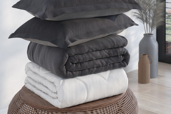 Image of a pile of bedding stacked on a round, wooden side table. The stack includes (from top to bottom): two Shadow/Dove Gray Pillow Shams, a Shadow/Dove Gray Duvet Cover, and a Duvet Insert all neatly folded