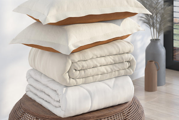 Image of a pile of bedding stacked on a round, wooden side table. The stack includes (from top to bottom): two reversible Ivory/Clay Pillow Shams, an ivory Duvet Cover, and a Duvet Insert all neatly folded