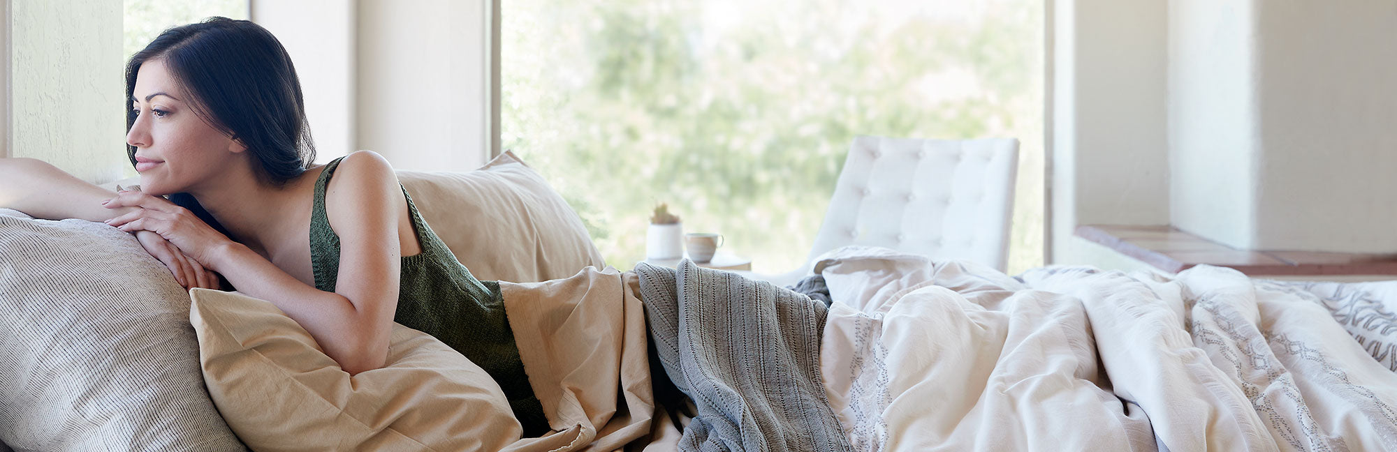 Image of a woman lying in bed, leaning over her pillows to look out the window with the Sonoran Bundle on her bed
