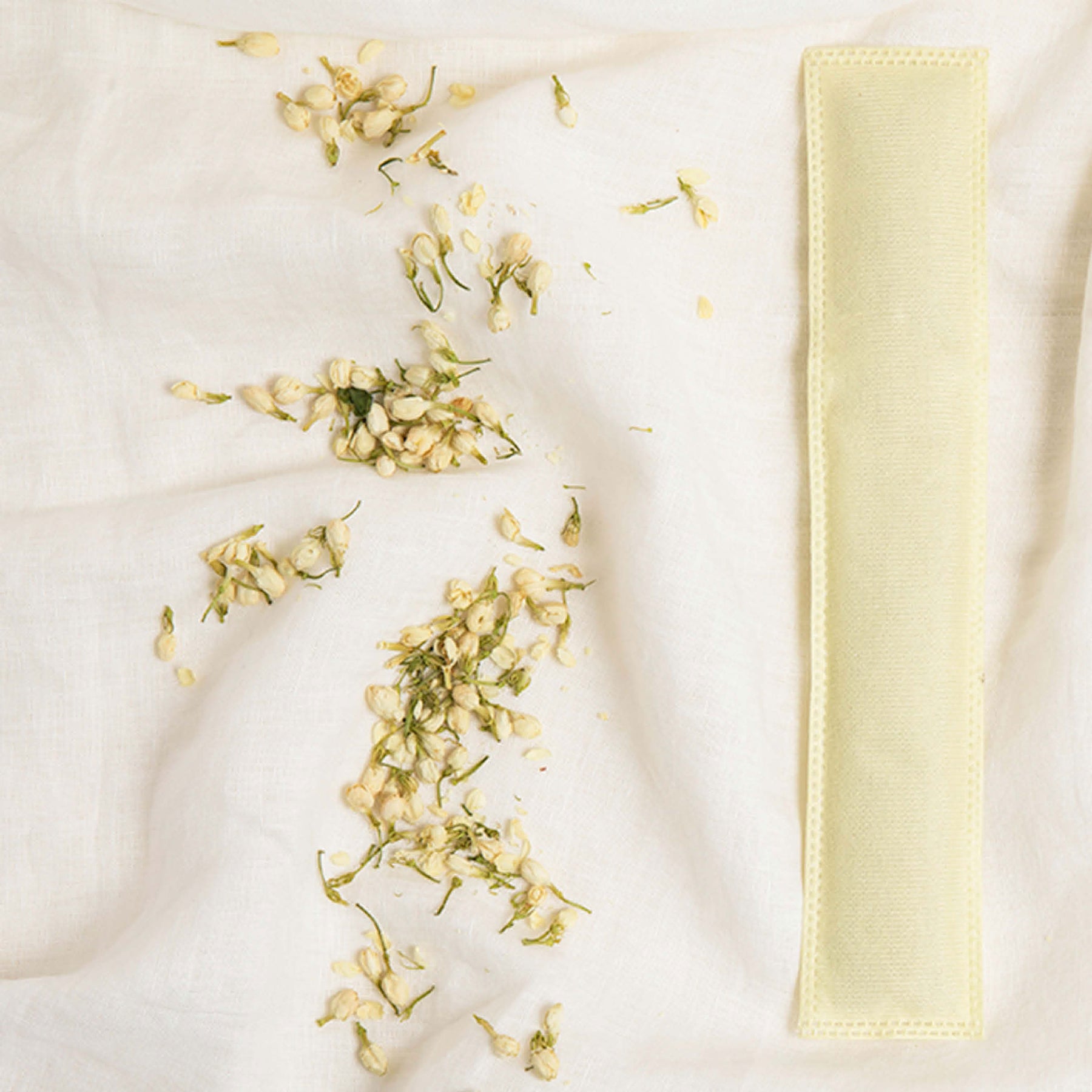 Image of a yellow Aromatherapy sachet with bits of jasmine flowers to the left of it on an off-white fabric background