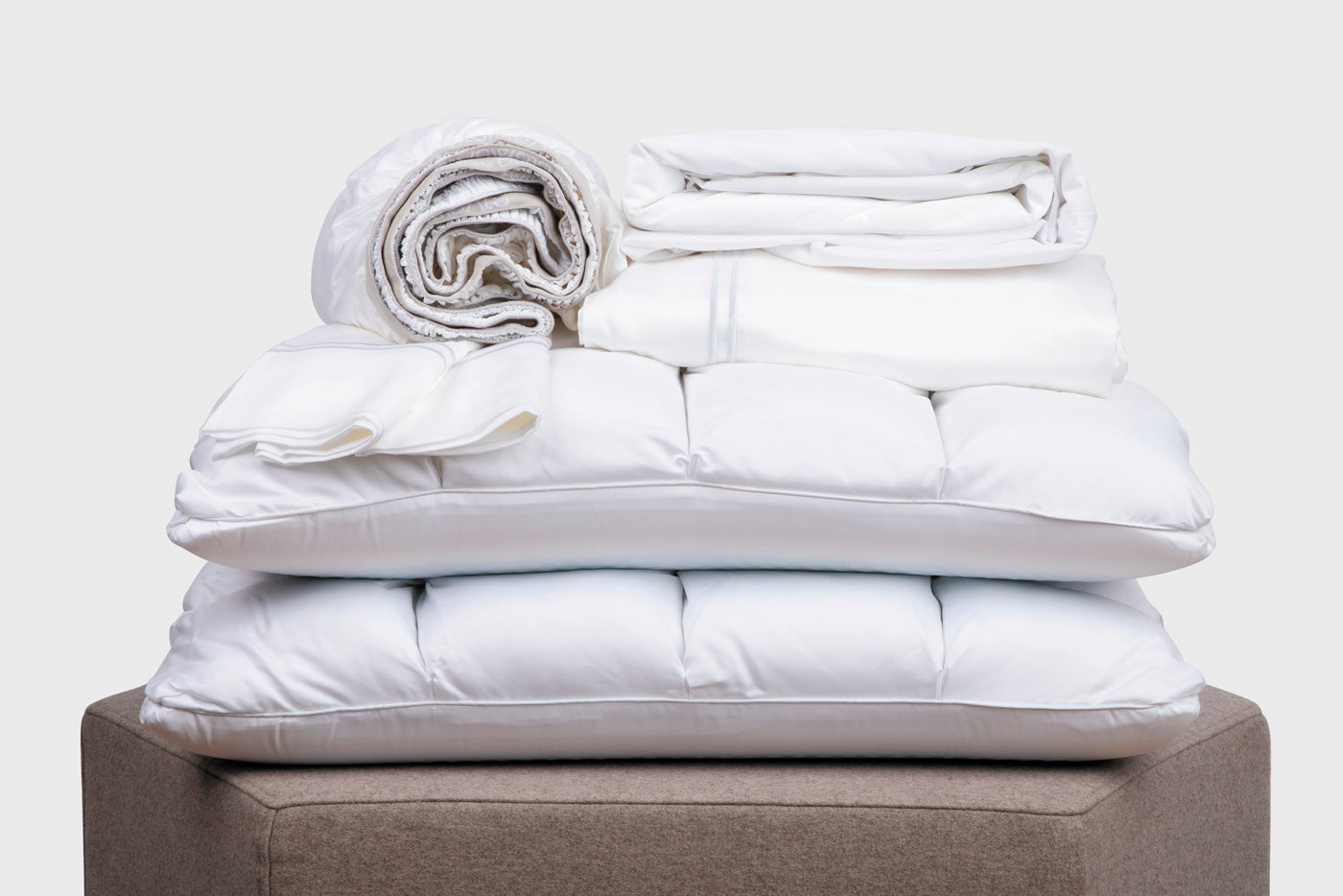 Image of a pile of all-white bedding on a taupe colored stool. The bedding includes (from top to bottom): a rolled white Refreshing TENCEL™ Lyocell fitted sheet, two white Refreshing TENCEL™ Lyocell Pillowcases, a neatly folded Cooling Mattress Protector, a neatly folded Refreshing TENCEL™ Lyocell flat sheet, and two SoftCell® Chill Pillows