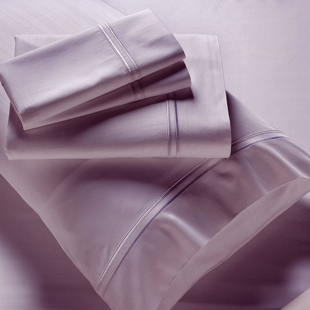 Image showcasing entire Lilac Bamboo Rayon Sheet Set. The image includes: a fitted sheet on the bed, a pillowcase on a pillow, a neatly folded flat sheet, and two neatly folded pillowcases.