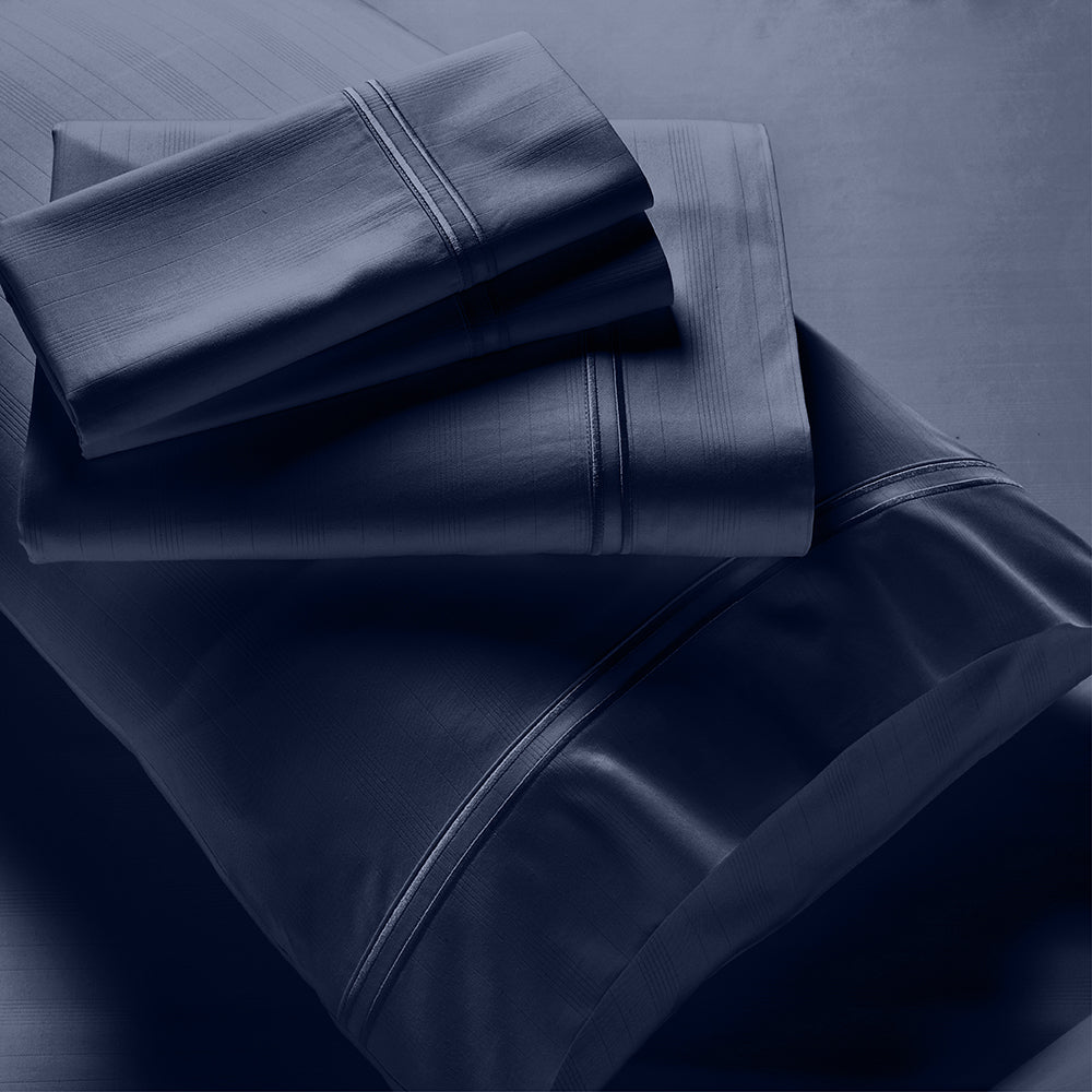 Image showcasing entire Midnight Bamboo Rayon Sheet Set. The image includes: a fitted sheet on the bed, a pillowcase on a pillow, a neatly folded flat sheet, and two neatly folded pillowcases.