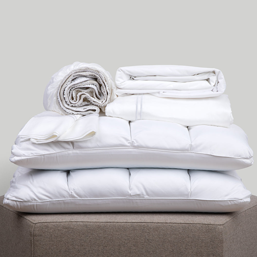 Image of a pile of all-white bedding on a taupe colored stool. The bedding includes (from top to bottom): a rolled white Refreshing TENCEL™ Lyocell fitted sheet, two white Refreshing TENCEL™ Lyocell Pillowcases, a neatly folded Cooling Mattress Protector, a neatly folded Refreshing TENCEL™ Lyocell flat sheet, and two SoftCell® Chill Pillows