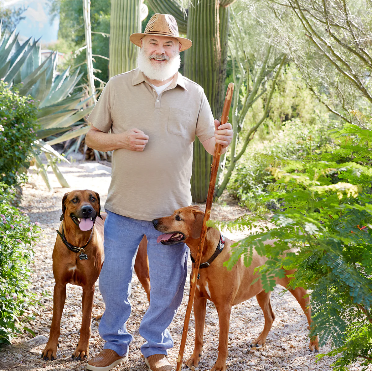 Image of Dr. Andrew Weil outside surrounded by cacti and other green plants with two Rhodesian Ridgeback dogs on either side of him