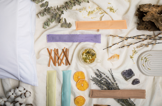 Image of 5 colored scented sachets and various plants that match each scent all laid out on an off-white fabric
