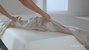 Video of a person making a bed with Ivory Bamboo Rayon Sheets. They start by tucking a fitted sheet on a mattress and showcasing the elastic band underneath. They then lay the extra-wide flat sheet over top. In the next shot, they are laying out the pillowcase (on a pillow) on top of a table to show the enveloping feature. 