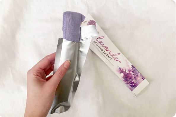 Image of a hand opening a purple Aromatherapy sachet from its silver packaging with a white fabric background