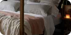 Image of a made bed with a dark background. The bed showcases pillows of various sizes and layered bedding. Items on the bed include: Kapok Pillows, White Blended Linen Sheets, Pinstripe Relaxed Hemp Pillowcases, a Pink Sandstone Wave Coverlet, and a reversible gray and white striped Heritage Quilt.