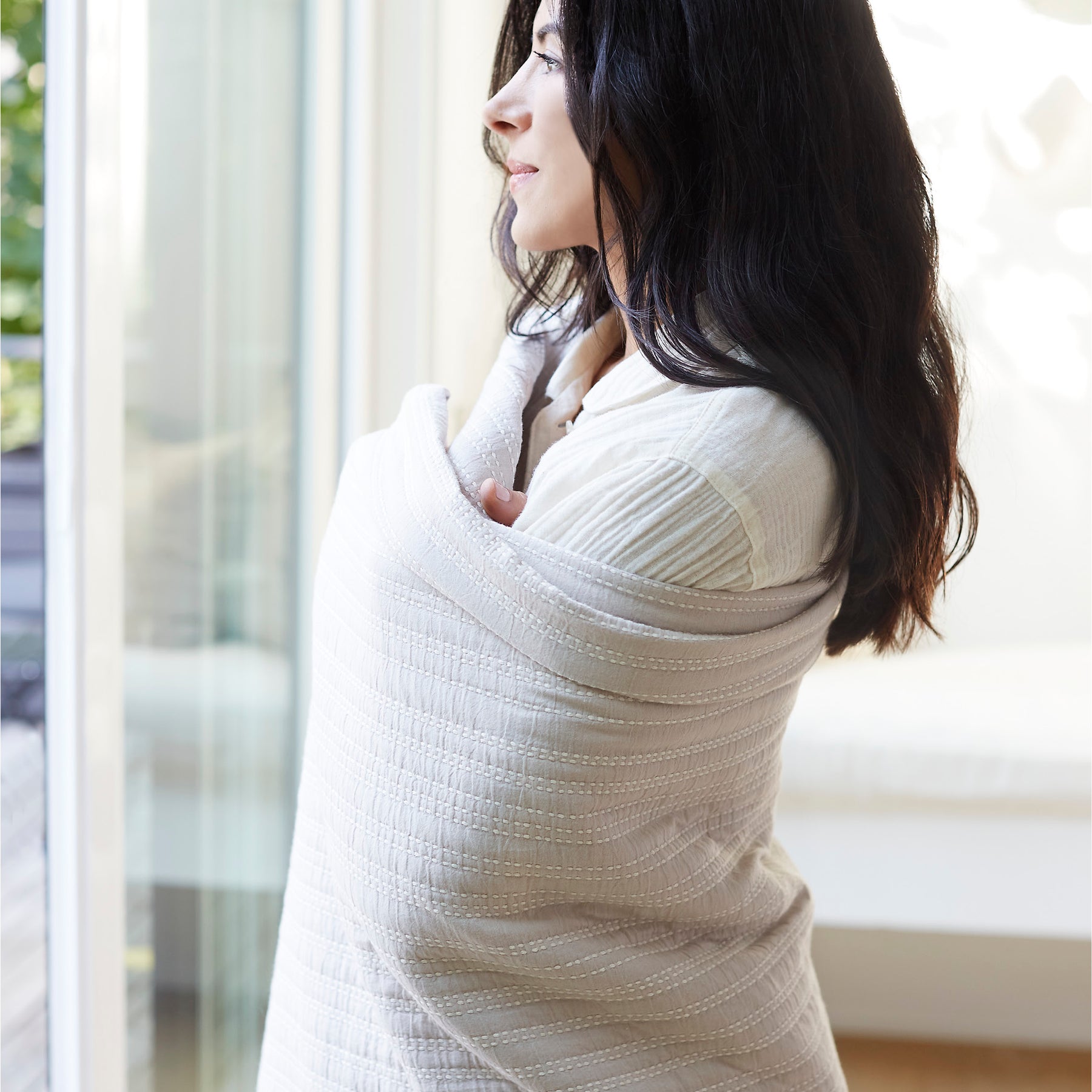 Image of a woman standing and staring out a window while wrapped up in an Oatmeal Everyday Cotton Coverlet