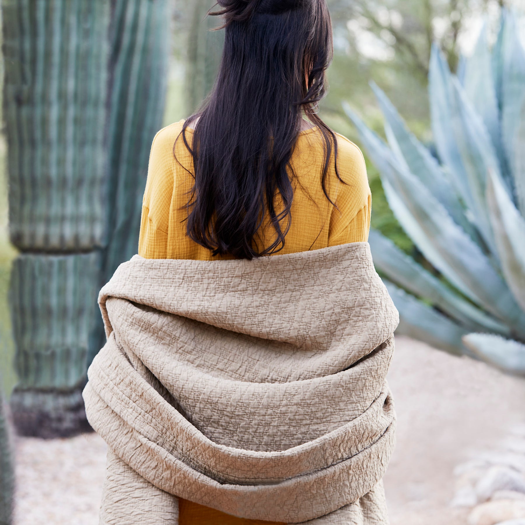 Image of a woman with long black hair and a yellow shirt standing outside by cacti. She is facing away, wrapped up in an Ochre Wave Coverlet.