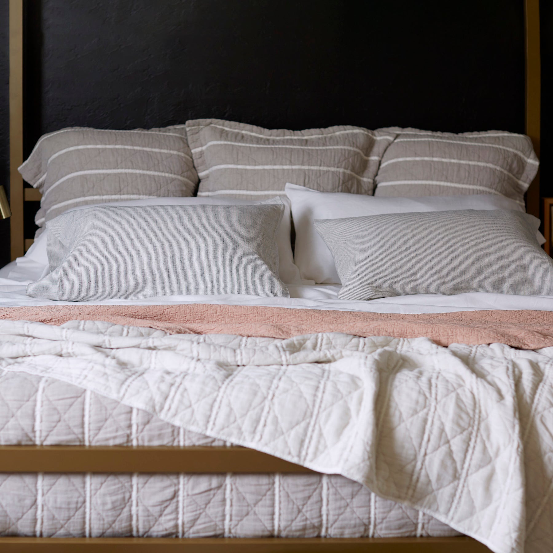Image of a made bed with a dark background. The bed showcases pillows of various sizes and layered bedding. Items on the bed include: Kapok Pillows, White Blended Linen Sheets, Pinstripe Relaxed Hemp Pillowcases, a Pink Sandstone Wave Coverlet, and a reversible gray and white striped Heritage Quilt.