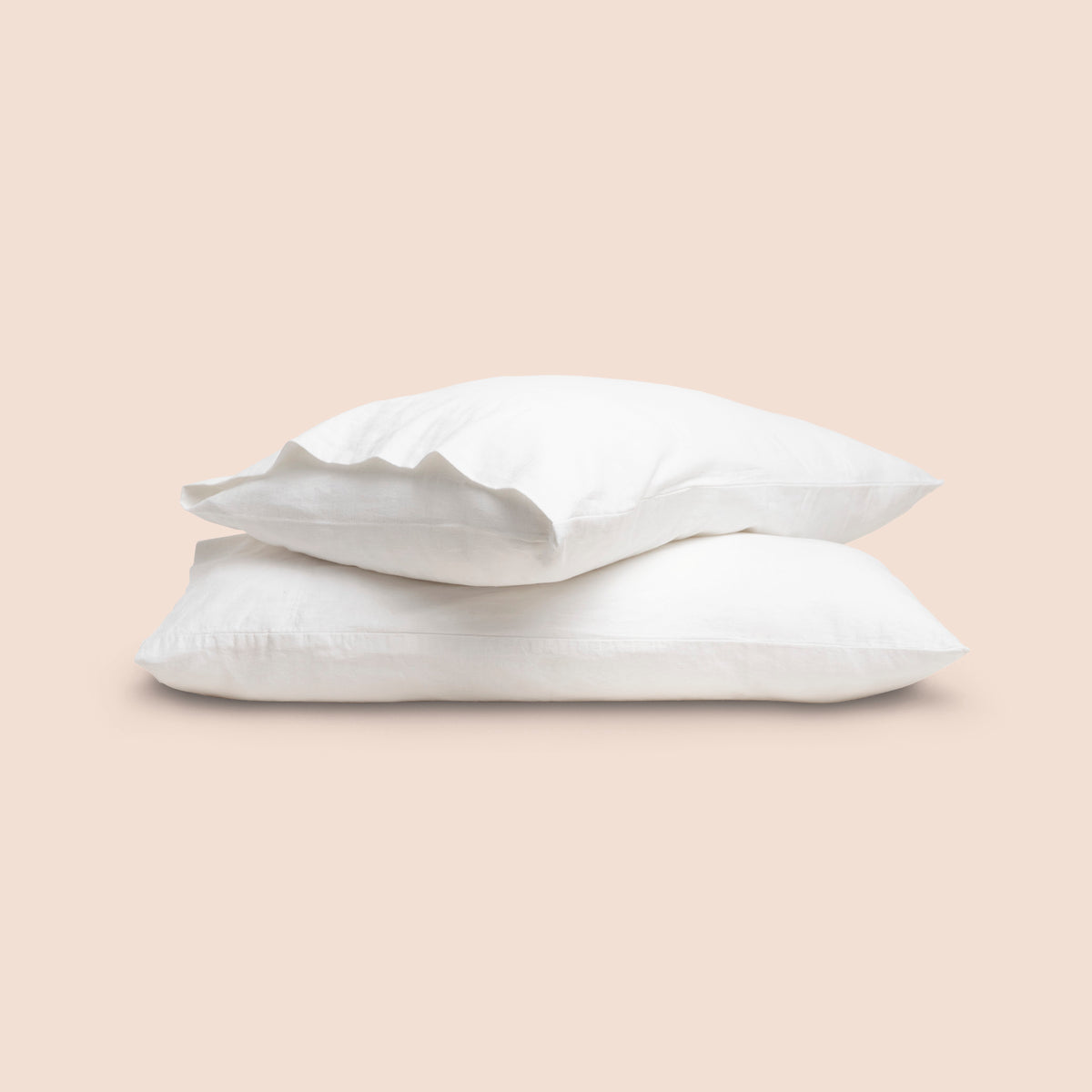 Image of two pillows with White Blended Linen Pillowcases stacked on top of each other on a light pink background. The top pillow is showcasing an enveloping feature. 