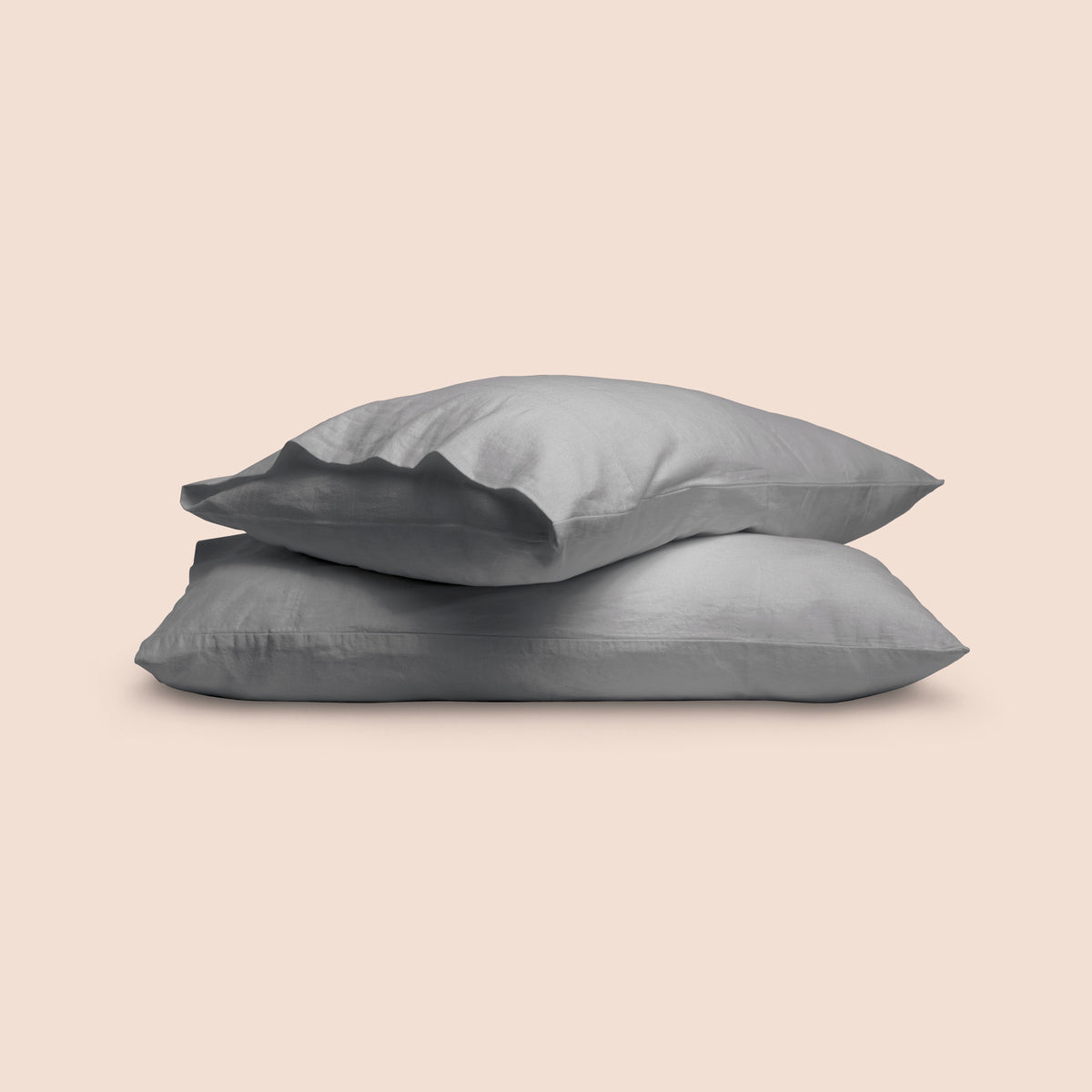 Image of two pillows with Stone Gray Blended Linen Pillowcases stacked on top of each other on a light pink background. The top pillow is showcasing an enveloping feature. 