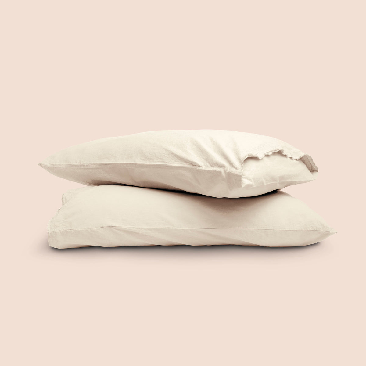 Image of two pillows with Ecru Garment Washed Percale pillowcases stacked on top of each other on a light pink background. The top pillow is showcasing an enveloping feature. 