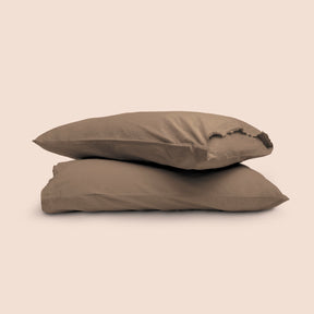 Image of two pillows with Desert Sand Garment Washed Percale pillowcases stacked on top of each other on a light pink background. The top pillow is showcasing an enveloping feature. 