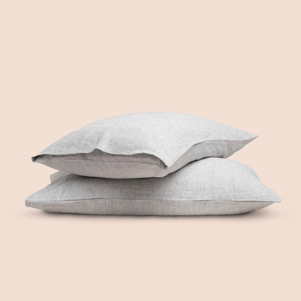 Image of two pillows with Pinstripe Relaxed Hemp pillowcases stacked on top of each other on a light pink background. The top pillow is showcasing an enveloping feature. 