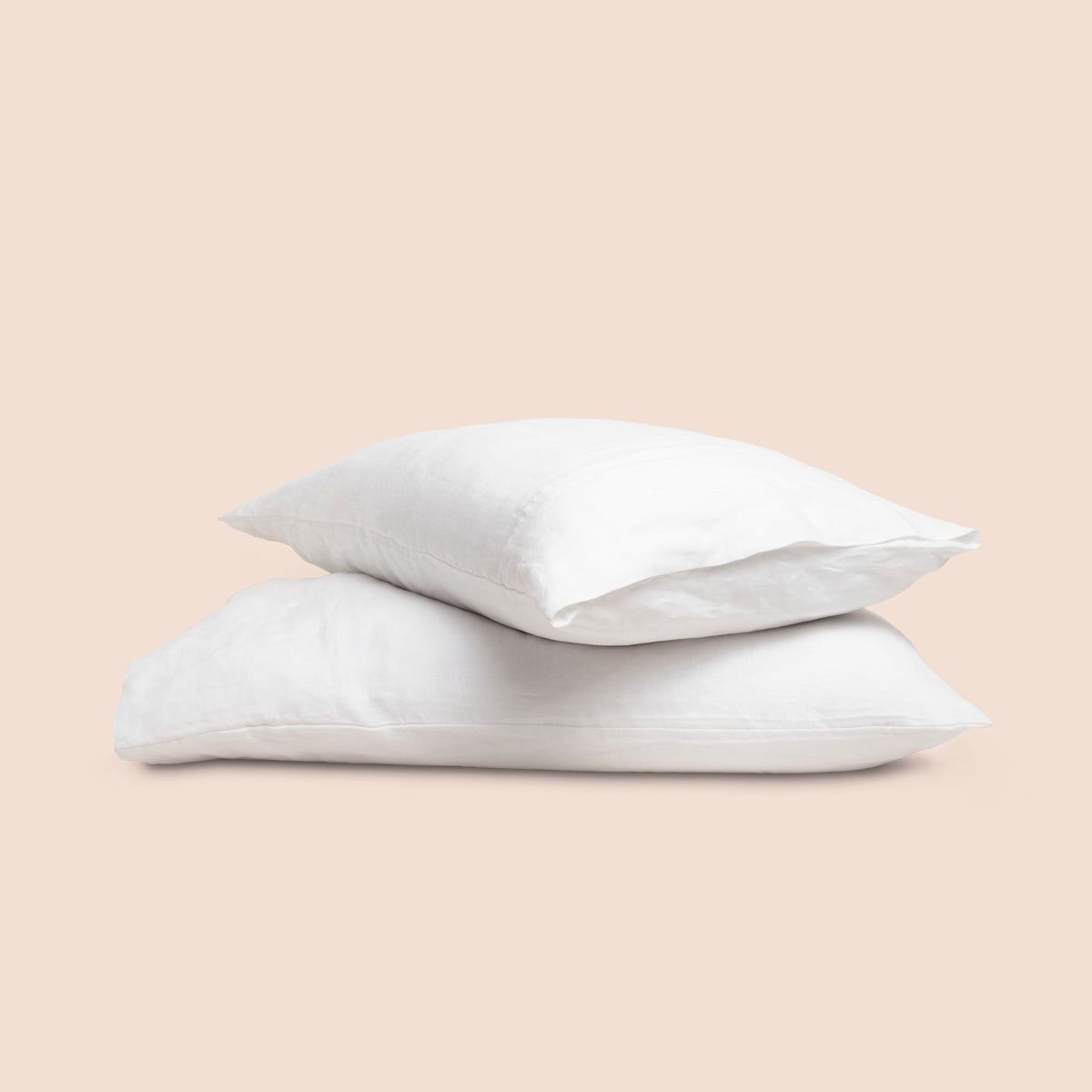 Image of two pillows with White Relaxed Hemp pillowcases stacked on top of each other on a light pink background. The top pillow is showcasing an enveloping feature. 